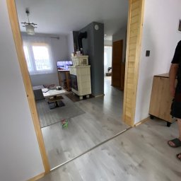 Zielpol 2.0 Dariusz Zieliński - Jasne wnętrze domu z drewnianą ramą drzwiową, widok na salon z kominkiem kaflowym i telewizorem. Podłoga z paneli imitujących drewno. Częściowo widoczna osoba.