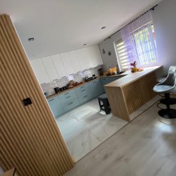 Zielpol 2.0 Dariusz Zieliński - Nowoczesna kuchnia z wyspą i drewnianą ścianką działową. Jasne fronty szafek, marmurowe płytki podłogowe i designerskie krzesła barowe. Ukończona realizacja.