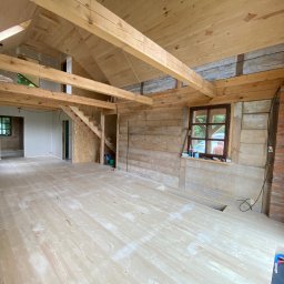 Zielpol 2.0 Dariusz Zieliński - Wnętrze domu w trakcie budowy z drewnianą konstrukcją, podłogą z desek i widocznymi elementami instalacji. Surowe wykończenie, okno z widokiem na zieleń.