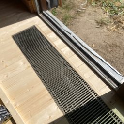 Zielpol 2.0 Dariusz Zieliński - Montaż kratki wentylacyjnej w drewnianej podłodze, widoczne narzędzia i elementy konstrukcyjne. Jasne drewno, metalowa kratka, częściowo widoczne buty.