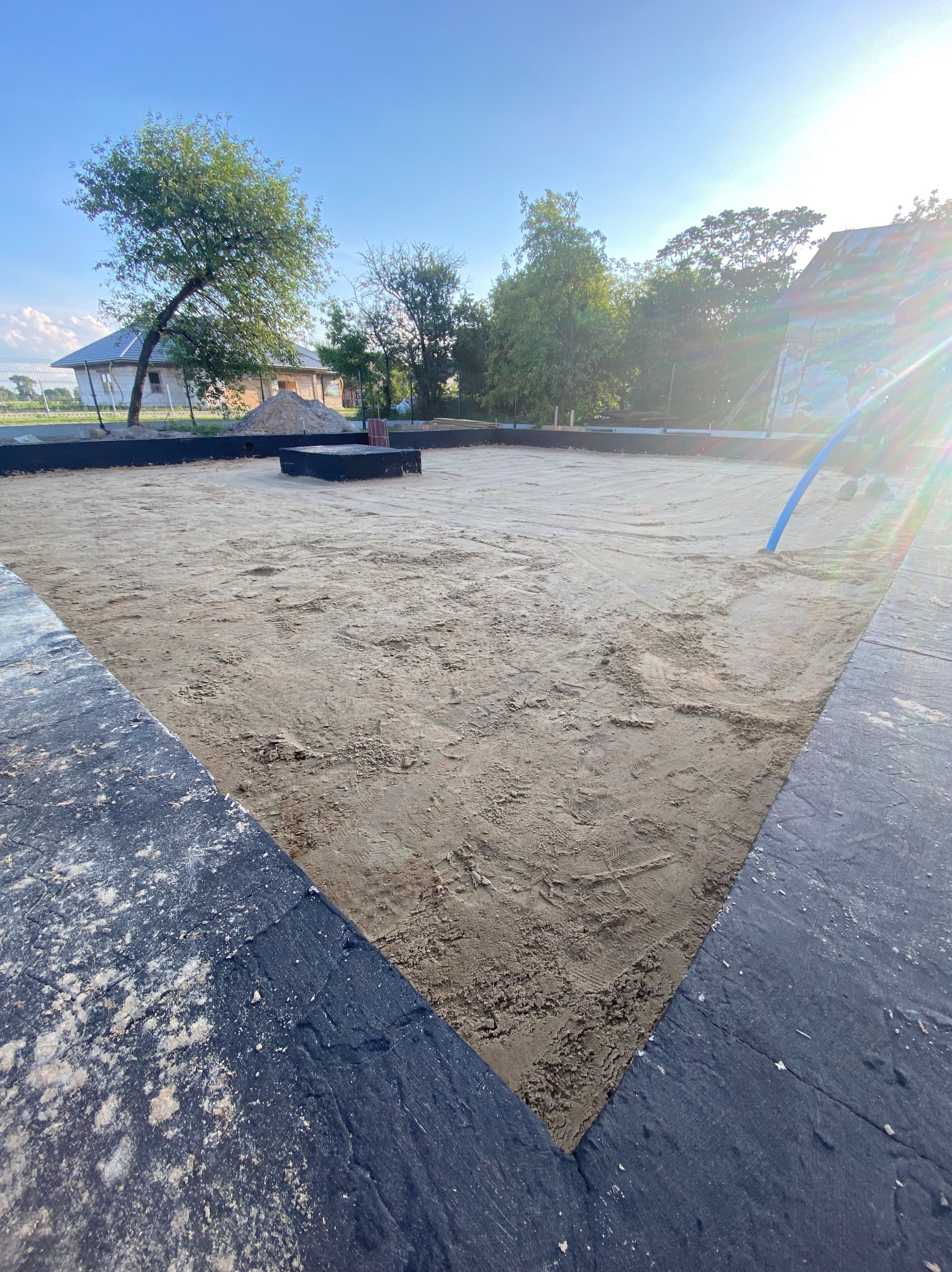 Przygotowany teren pod budowę domu z widocznymi fundamentami i piaskiem. W tle dom w trakcie budowy i zieleń. Słoneczny dzień.