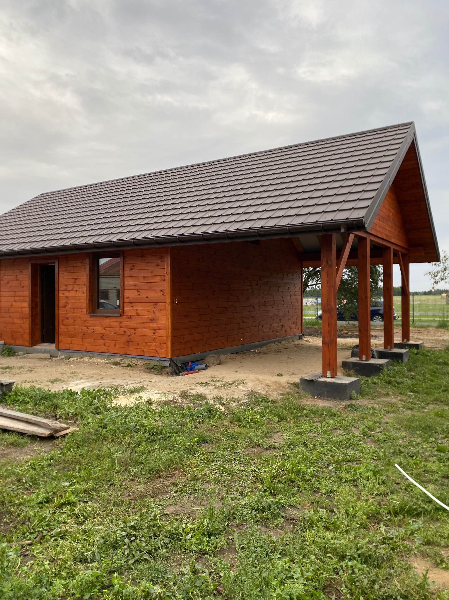 Drewniany dom szkieletowy z brązowym dachem, widok z zewnątrz, częściowo zadaszony taras. Widoczne elementy konstrukcyjne i wykończeniowe. Otoczenie: trawa, ziemia.