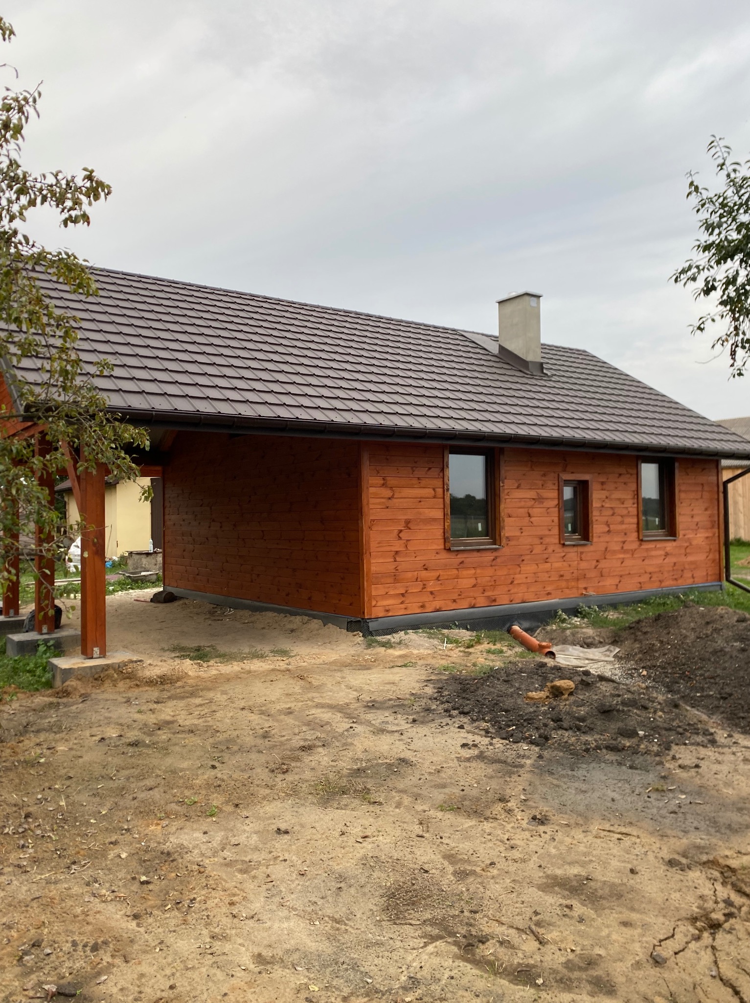 Drewniany dom z brązowym dachem, widok z boku, zadaszone wejście. Otoczenie w trakcie prac ziemnych, rura odpływowa. Pochmurne niebo w tle.