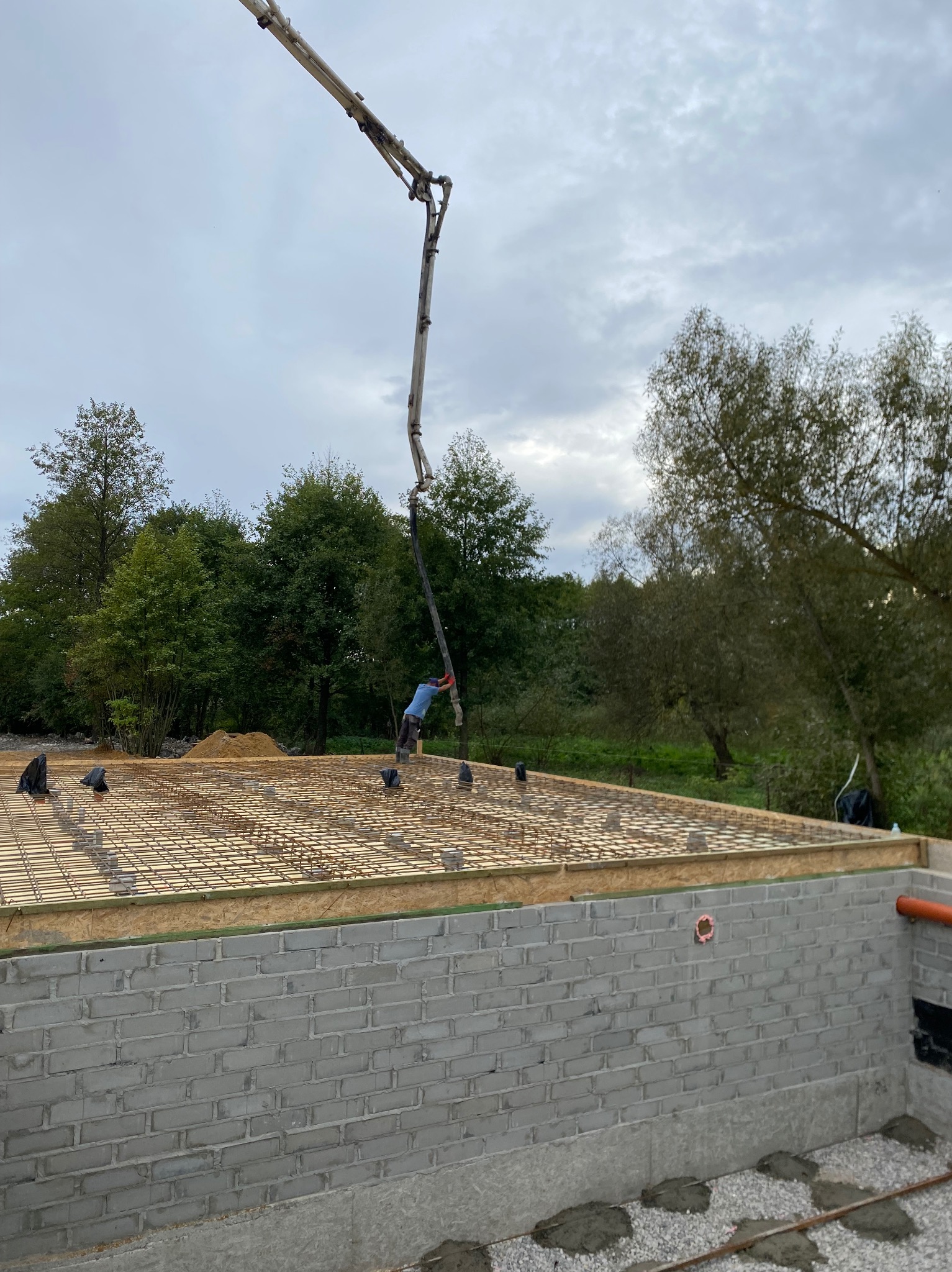 Wylewka fundamentu domu z betonomieszarki. Widoczne zbrojenie, szalunek i ściany fundamentowe z bloczków. W tle drzewa i pochmurne niebo.