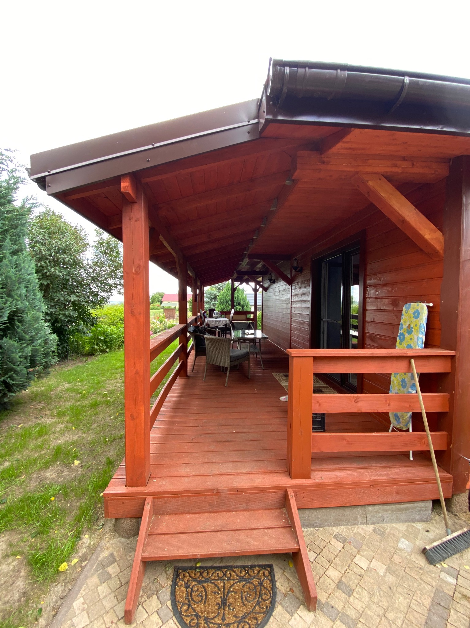 Drewniana altana z zadaszeniem, widok z zewnątrz. Czerwono-brązowa konstrukcja, schody wejściowe, meble ogrodowe w tle. Ujęcie z niskiej perspektywy.