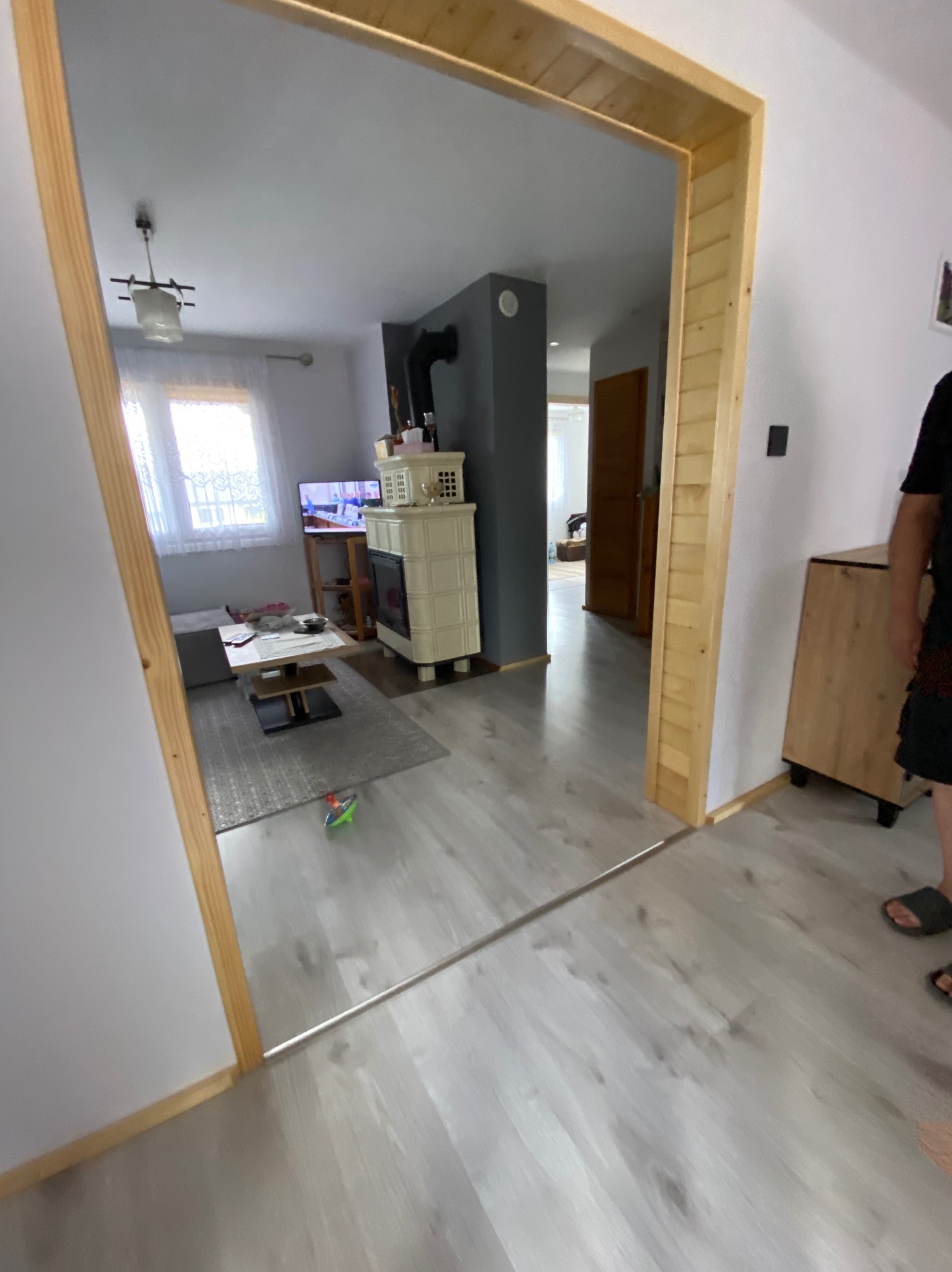 Jasne wnętrze domu z drewnianą ramą drzwiową, widok na salon z kominkiem kaflowym i telewizorem. Podłoga z paneli imitujących drewno. Częściowo widoczna osoba.