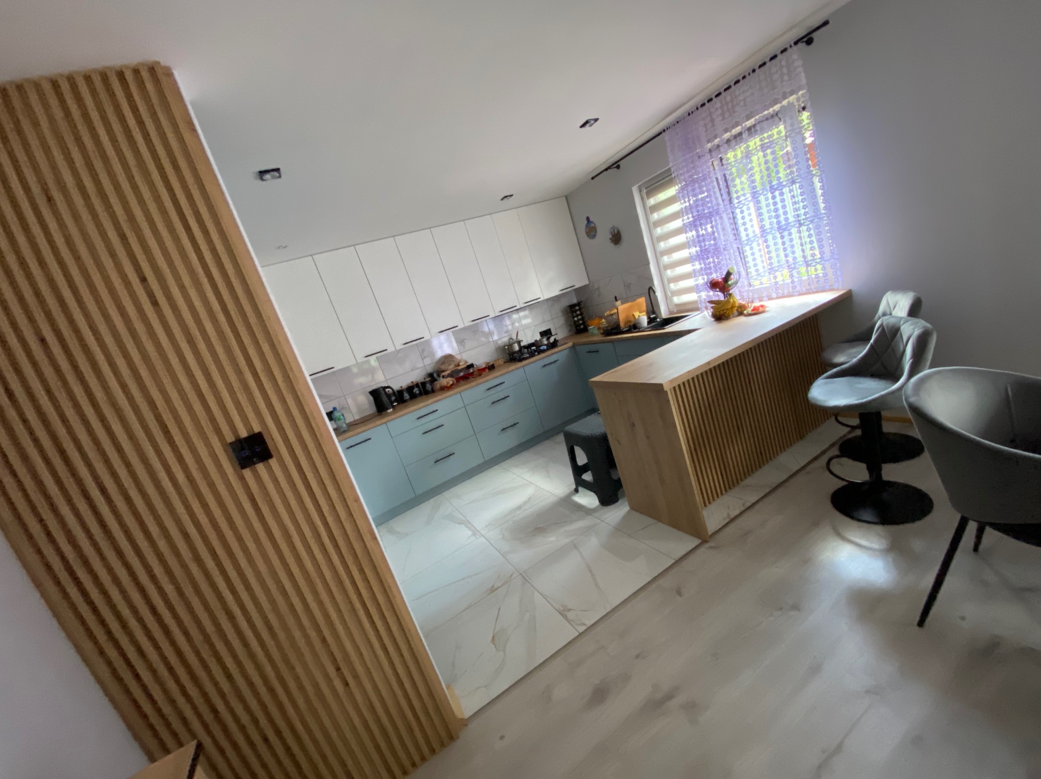 Nowoczesna kuchnia z wyspą i drewnianą ścianką działową. Jasne fronty szafek, marmurowe płytki podłogowe i designerskie krzesła barowe. Ukończona realizacja.