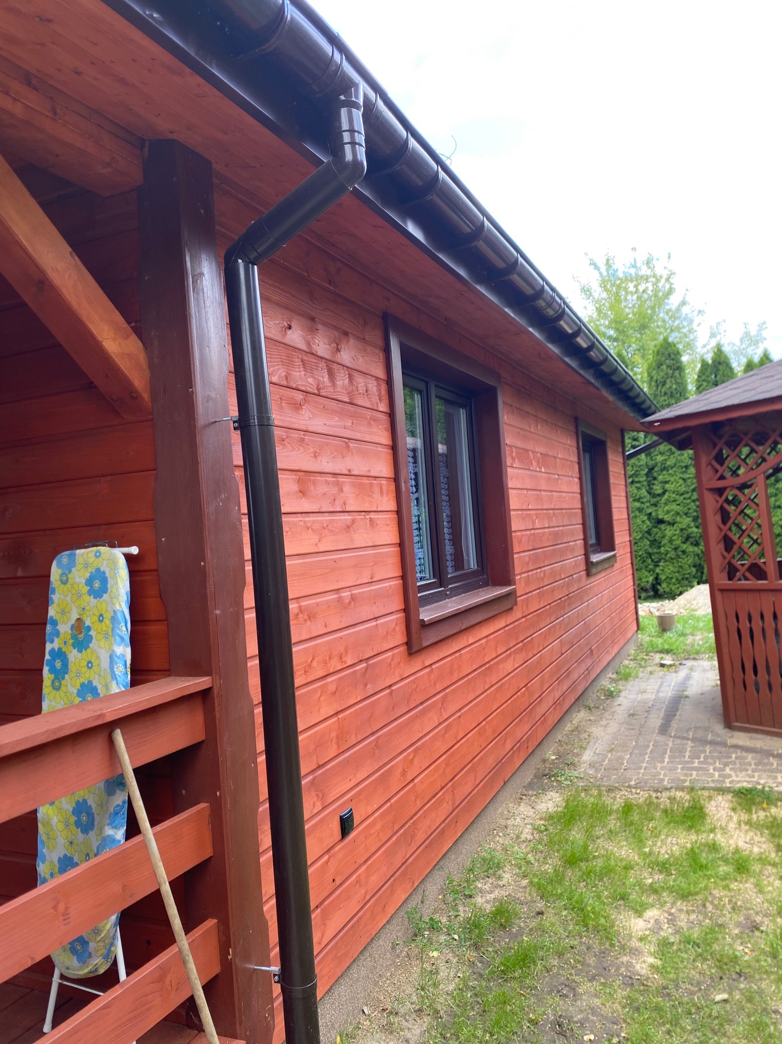 Czerwony dom z drewnianą elewacją i brązowymi rynnami. Widoczna altana w tle i deska do prasowania na balkonie. Ujęcie z perspektywy.