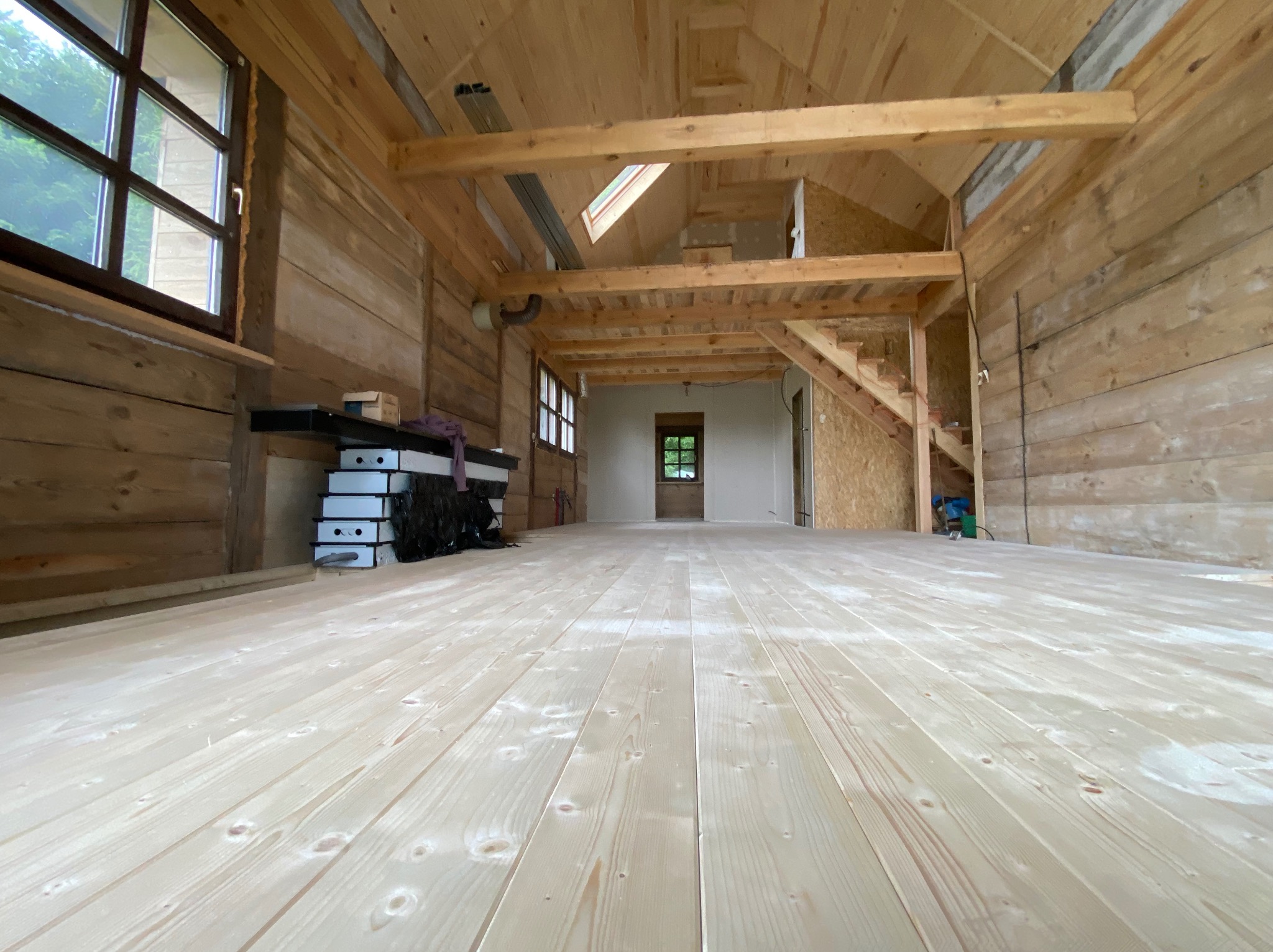 Wnętrze domu w trakcie budowy z drewnianą konstrukcją, podłogą z jasnego drewna i schodami. Widoczne okna i elementy instalacji.