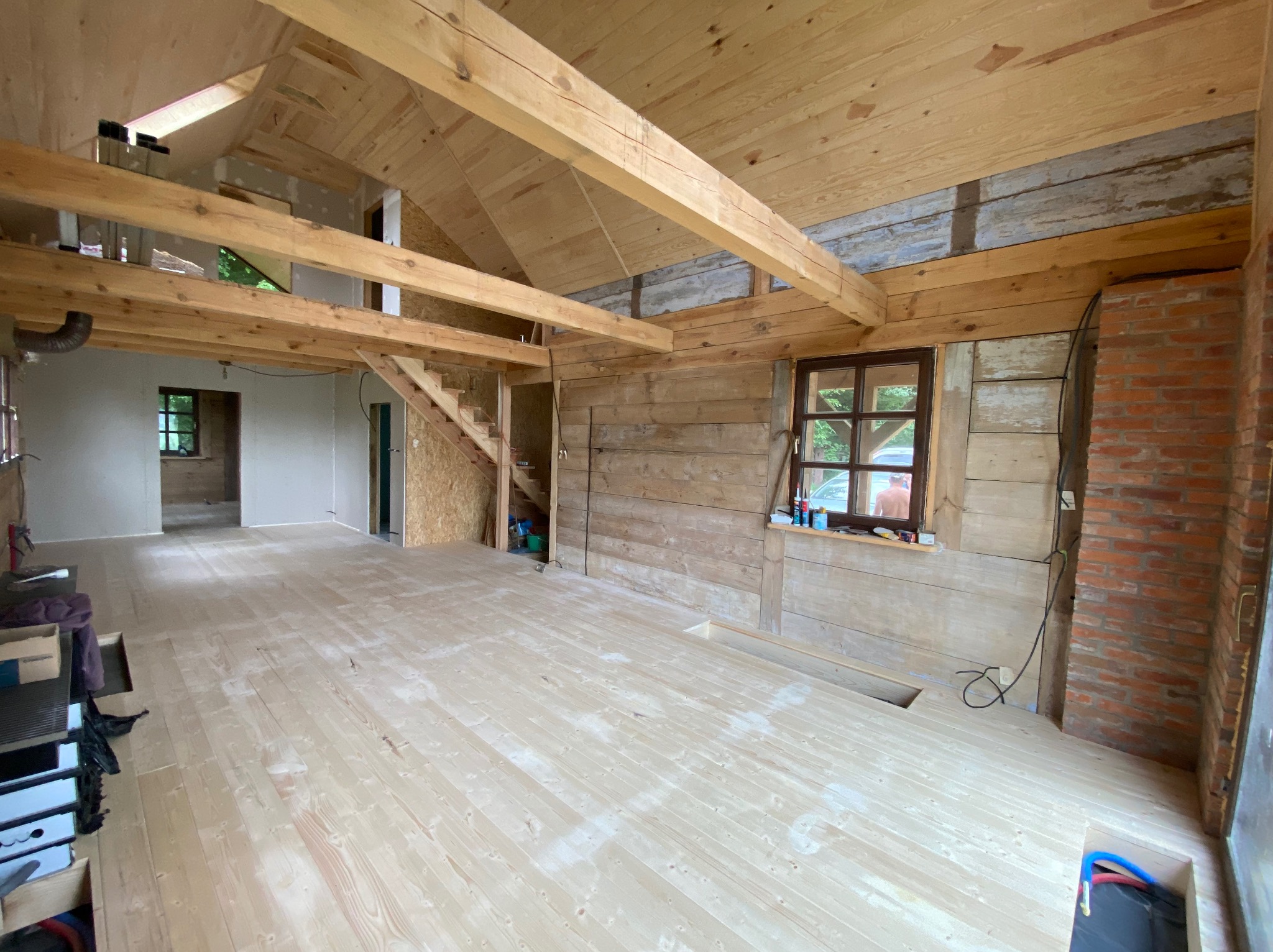 Wnętrze domu w trakcie budowy z drewnianą konstrukcją, podłogą z desek i widocznymi elementami instalacji. Surowe wykończenie, okno z widokiem na zieleń.