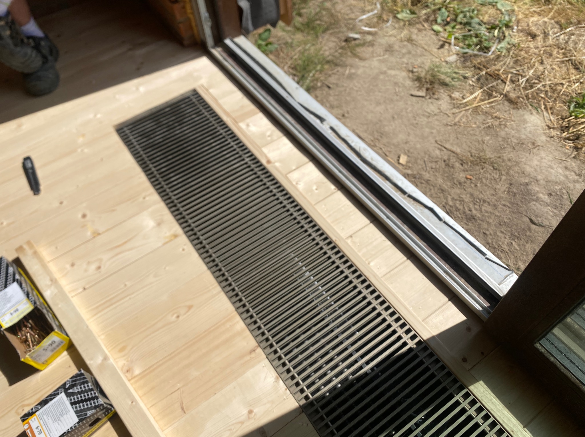 Montaż kratki wentylacyjnej w drewnianej podłodze, widoczne narzędzia i elementy konstrukcyjne. Jasne drewno, metalowa kratka, częściowo widoczne buty.