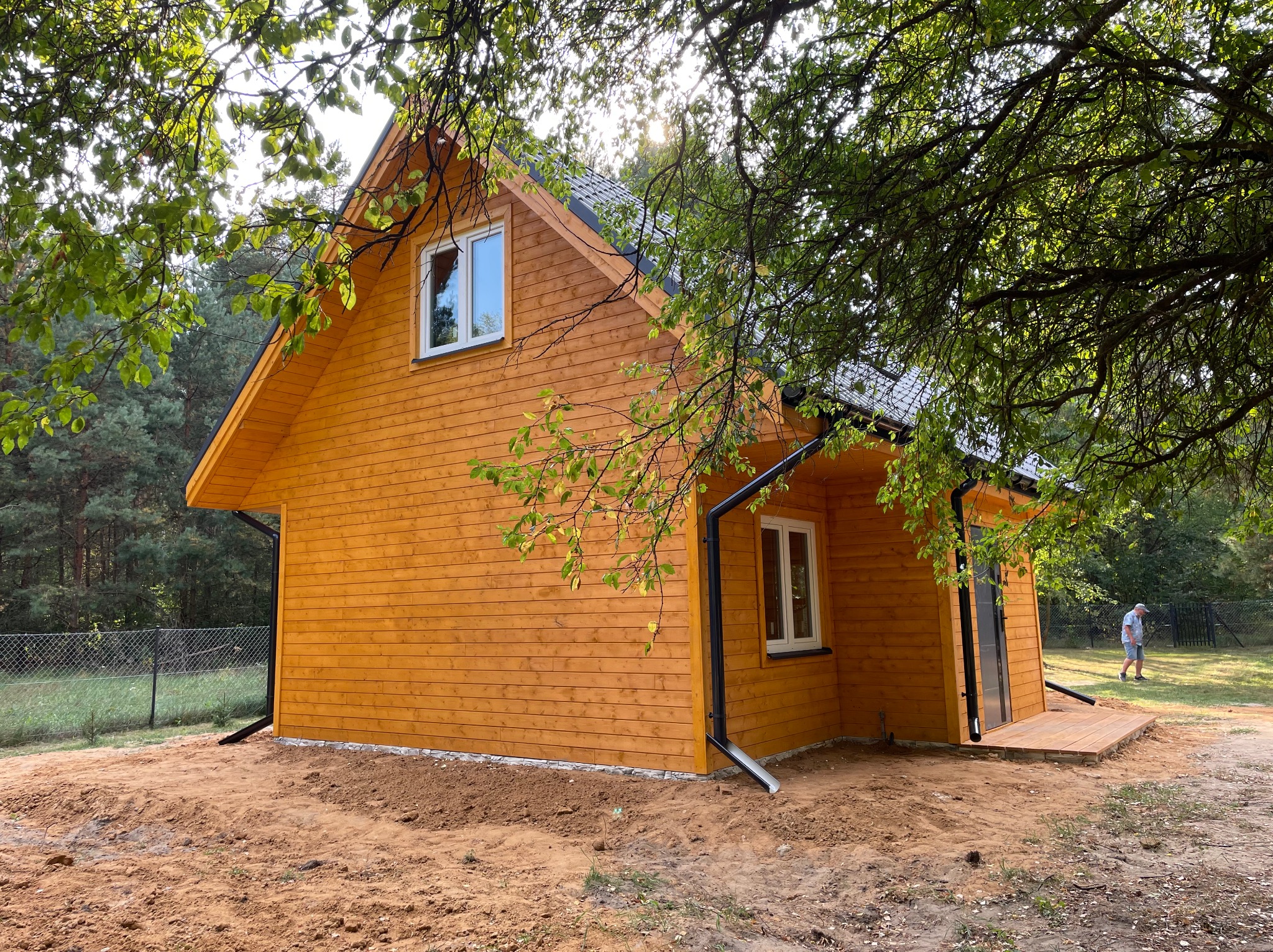 Nowy, drewniany dom szkieletowy z czarnym dachem i białymi oknami, otoczony drzewami i przygotowanym terenem. Widoczny fragment tarasu i rynny.