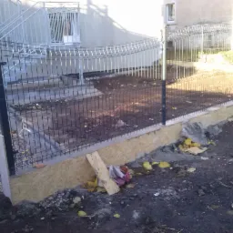 Nowy, metalowy płot panelowy z podmurówką z płyty OSB, zamontowany przy budynku. Widoczne schody z metalową barierką. Teren wokół ogrodzenia niezagospodarowany.