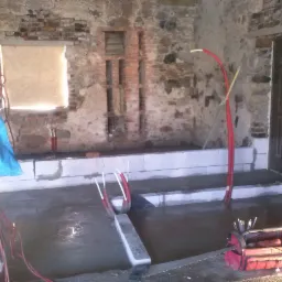 Wnętrze w trakcie prac murarskich: surowe ściany z cegły, wylana posadzka, widoczne instalacje i narzędzia w czerwonej torbie. Okno częściowo zasłonięte.