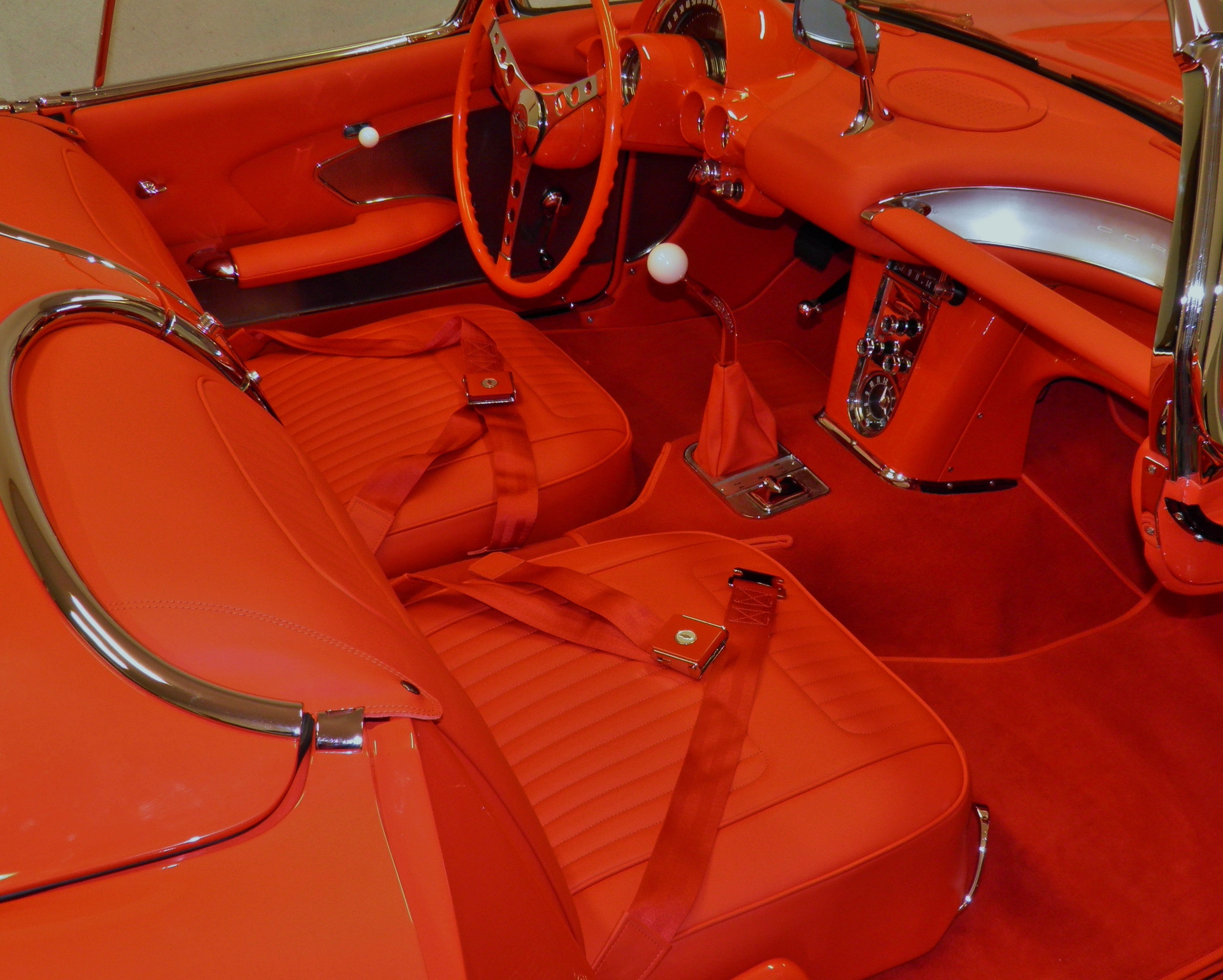 Wnętrze klasycznego, czerwonego samochodu sportowego z chromowanymi detalami. Fotele z pasami, deska rozdzielcza z zegarami i kierownica w stylu retro. Całość w odcieniach czerwieni.