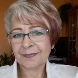 Portret kobiety w okularach, z krótkimi blond włosami z czerwonymi refleksami, ubrana w białą marynarkę, na tle pomarańczowej ściany i częściowo widocznych mebli.