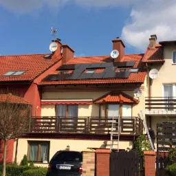 Czerwony dach z panelami słonecznymi w zabudowie szeregowej. Na dachu anteny satelitarne i mała turbina wiatrowa. Ustawiona drabina. Słoneczny dzień.