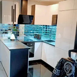 Nowoczesna kuchnia z białymi błyszczącymi szafkami, szklanym backsplashiem w odcieniach błękitu i czarnymi elementami, widoczne lampy wiszące i kurtka na krześle.