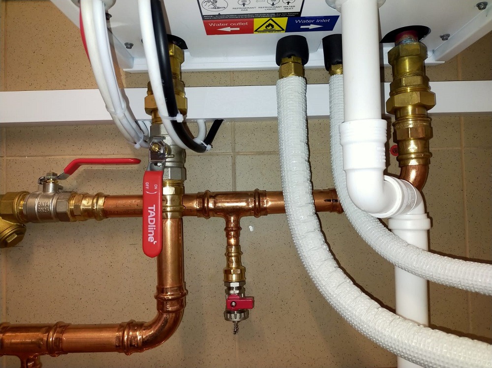 Kompleksowe rury miedziane i białe instalacje pompy ciepła z zaworami, widoczne złącza i izolowane przewody, na tle jasnych płytek.