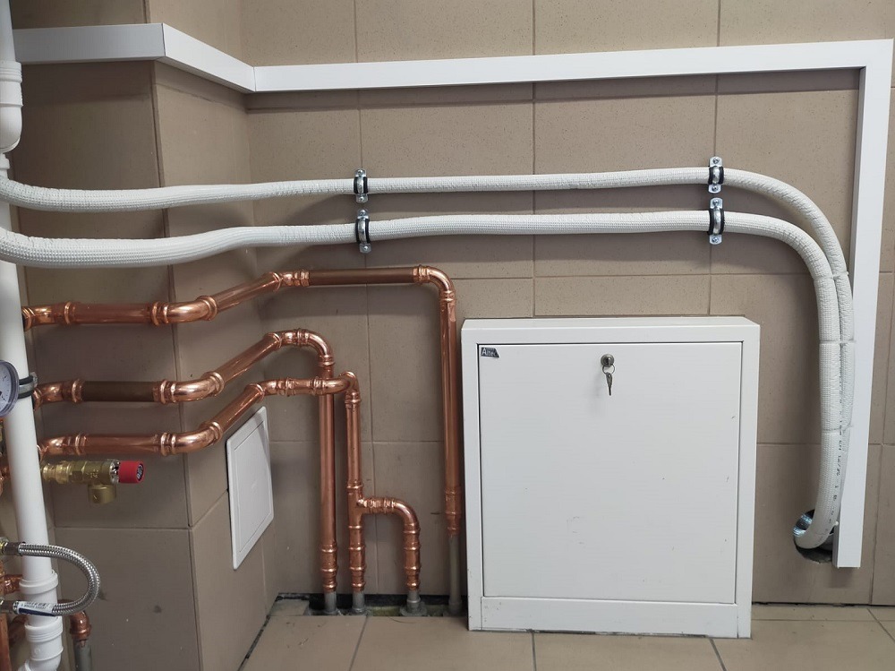 Miedziane rury instalacji grzewczej, białe rury izolowane i szafka z kluczem na tle beżowych płytek. Element instalacji pompy ciepła w pomieszczeniu.