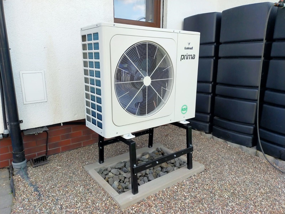 Biała pompa ciepła Galmet Prima, zamontowana na czarnym stelażu, na tle ściany budynku i zbiorników na wodę. Widoczny certyfikat R32.