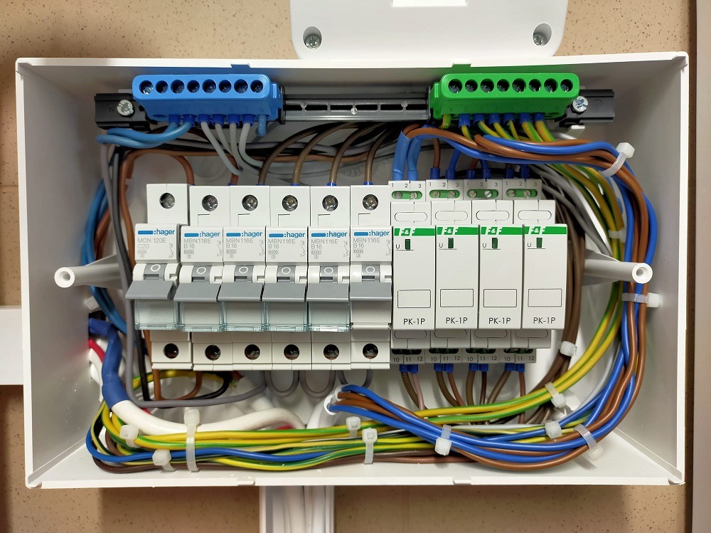 Otwarta skrzynka z bezpiecznikami Hager, widoczne kolorowe przewody, listwy zaciskowe (niebieska i zielona) oraz zabezpieczenia przepięciowe PK-1P. Estetyczne ułożenie przewodów.