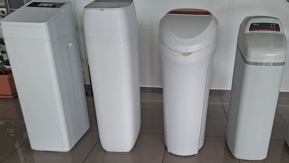 Cztery białe zmiękczacze wody różnych modeli, ustawione obok siebie na podłodze z płytek, prezentowane w jasnym oświetleniu wnętrza.