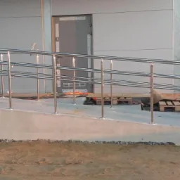 Zewnętrzna pochylnia z metalowymi barierkami ze stali nierdzewnej prowadząca do budynku z szarymi drzwiami, w tle widoczne drzewa.