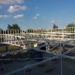 Konstrukcja stalowa budynku w trakcie budowy, zabezpieczona białym malowaniem, widoczna z góry. Na drugim planie zabudowa jednorodzinna i błękitne niebo z chmurami.