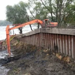 Pomarańczowa koparka przy brzegu rzeki, prowadząca prace ziemne. W tle woda i drzewa. Widoczne metalowe zabezpieczenia brzegu.