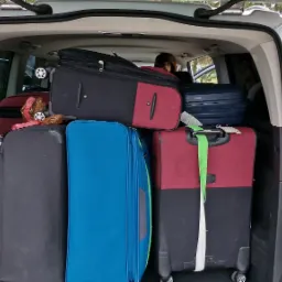 Bagażnik srebrnego vana wypełniony kolorowymi walizkami różnych rozmiarów, gotowy do podróży. Widoczny fragment siedzenia i osoba wewnątrz pojazdu.