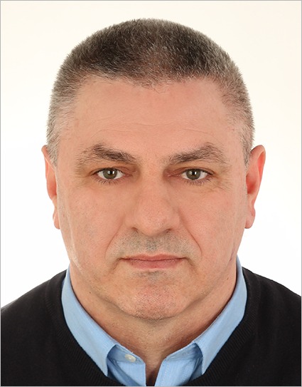 Portret mężczyzny w niebieskiej koszuli i czarnym swetrze na jasnym tle.