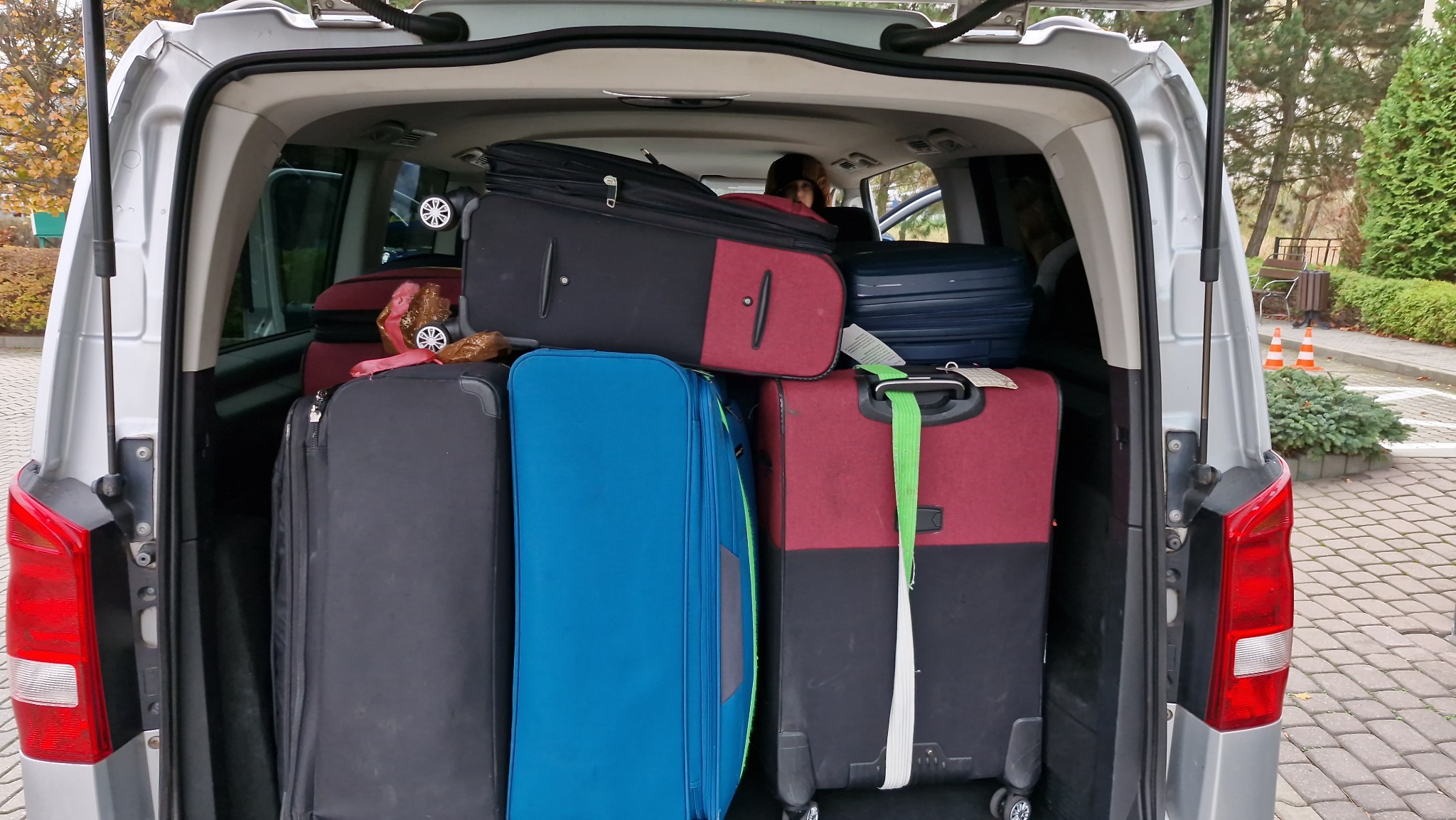 Bagażnik srebrnego vana wypełniony kolorowymi walizkami różnych rozmiarów, gotowy do podróży. Widoczny fragment siedzenia i osoba wewnątrz pojazdu.