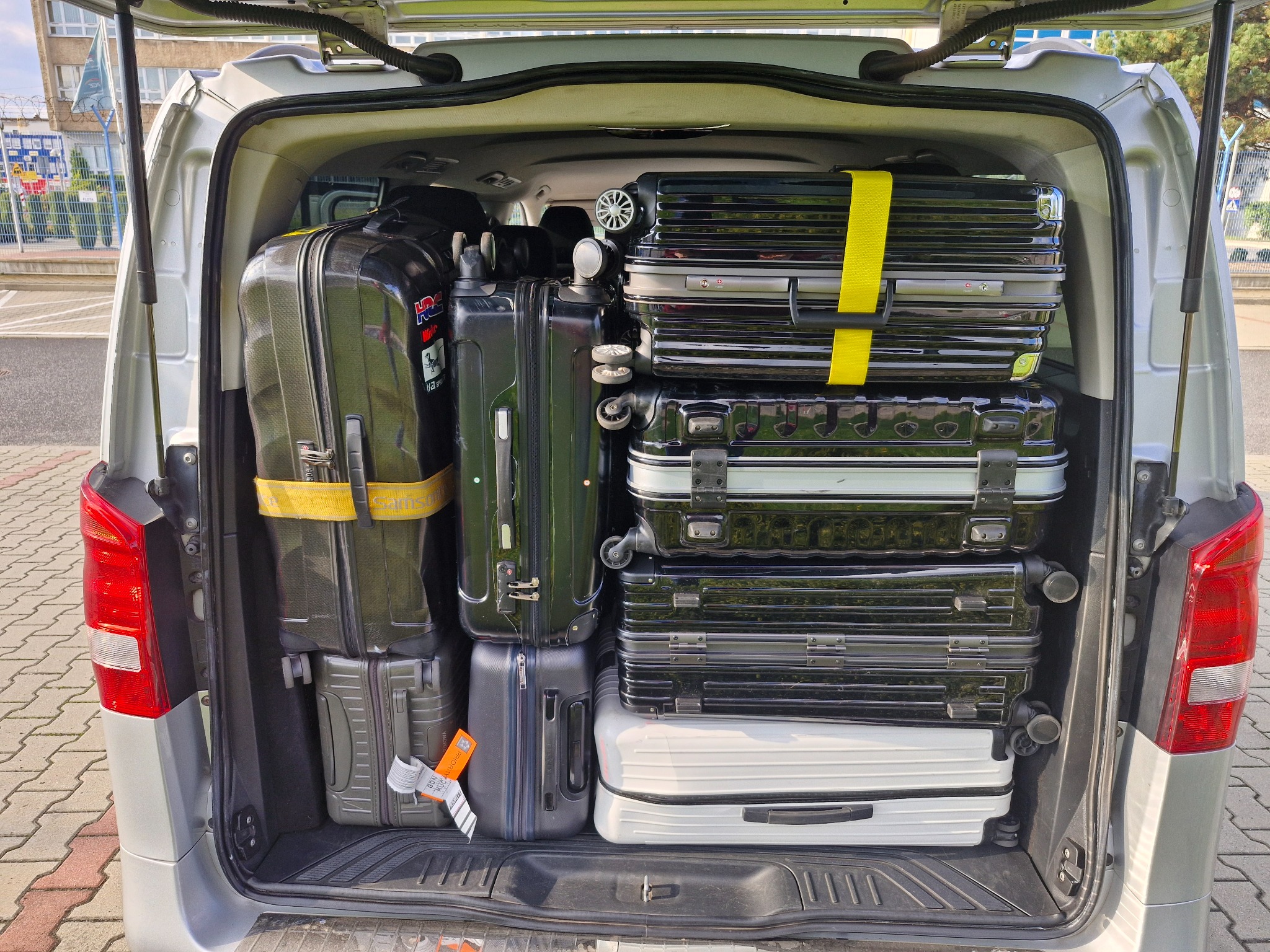 Bagażnik srebrnego vana wypełniony po brzegi różnymi walizkami, niektóre zabezpieczone żółtymi pasami. Widoczne etykiety bagażowe i fragment budynku w tle.