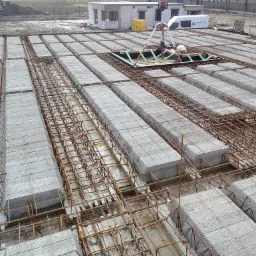 Szczegółowy widok z góry na strop Teriva z betonowymi pustakami i siatką zbrojeniową, otwór w stropie zabezpieczony drewnianą konstrukcją, w tle budynek w stanie surowym otwartym i pojazd dostawczy.