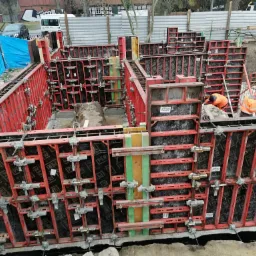 Szalunek fundamentów z czerwonymi ramami i czarną wyściółką, widoczni dwaj pracownicy w pomarańczowych kurtkach, przygotowujący zbrojenie w wykopie.