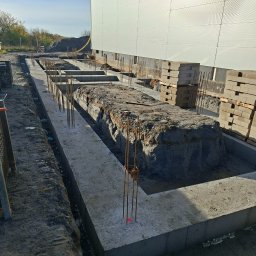 Widrog - Wykop pod fundamenty z częściowo wylanym betonem i wystającymi prętami zbrojeniowymi. W tle ściana budynku i palety z bloczkami.