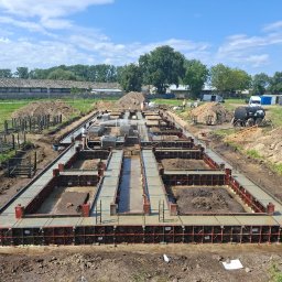 Widrog - Zalane betonem szalunki fundamentowe na budowie domu jednorodzinnego, widok z góry. W tle budynek gospodarczy i drzewa, błękitne niebo.