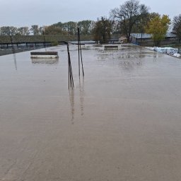 Widrog - Świeżo wylany, mokry beton na dużej powierzchni z wystającymi prętami zbrojeniowymi. W tle budynki gospodarcze i drzewa pod pochmurnym niebem.