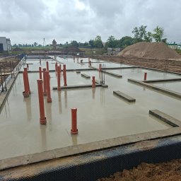 Widrog - Zalany fundament z pionowymi rurami kanalizacyjnymi na budowie. Widoczne zbrojenia, budynek w tle i hałda piasku. Pochmurne niebo.