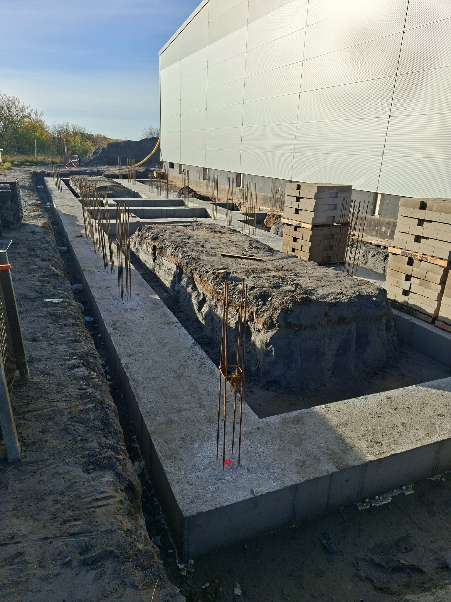 Wykop pod fundamenty z częściowo wylanym betonem i wystającymi prętami zbrojeniowymi. W tle ściana budynku i palety z bloczkami.