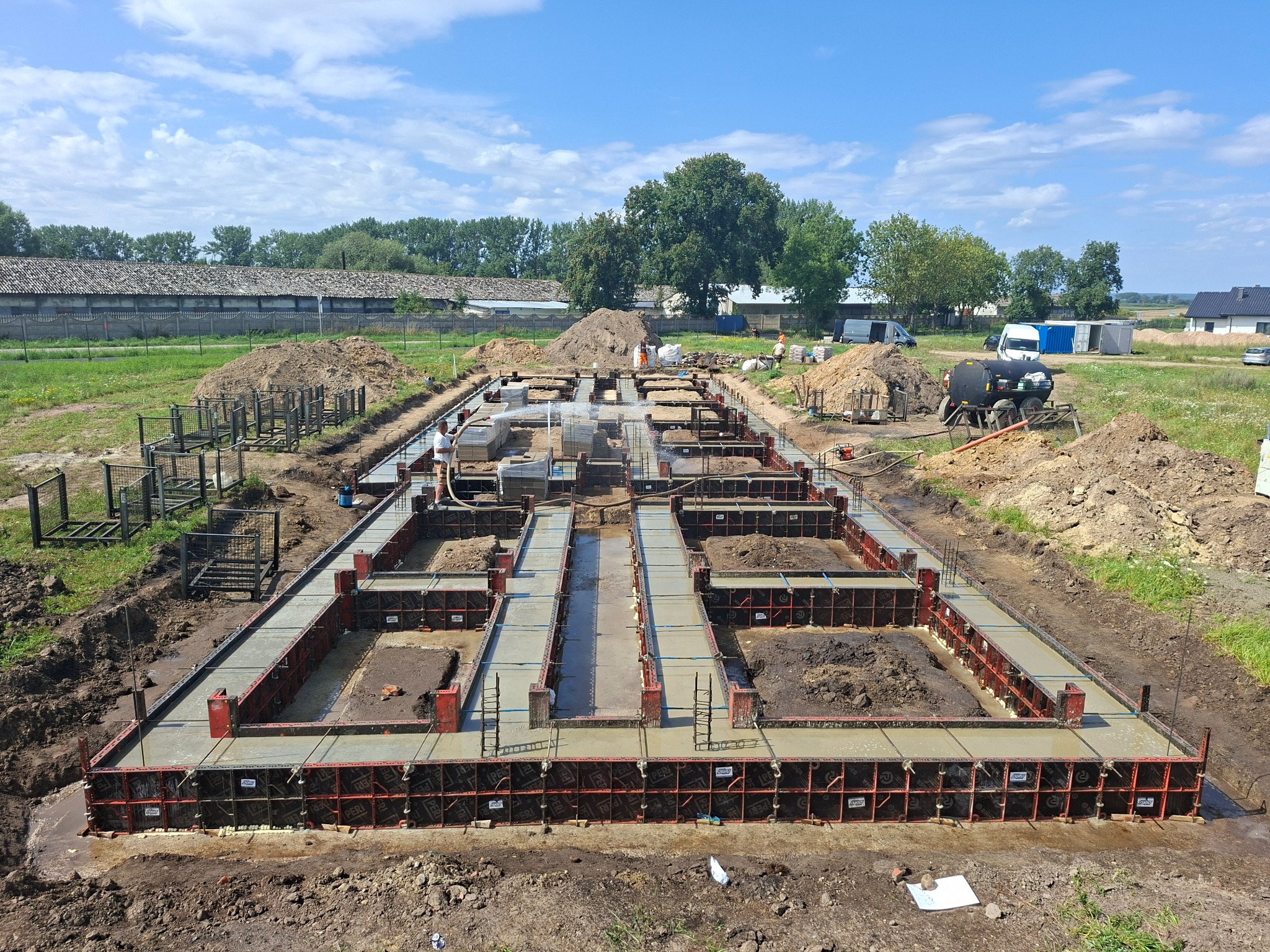Zalane betonem szalunki fundamentowe na budowie domu jednorodzinnego, widok z góry. W tle budynek gospodarczy i drzewa, błękitne niebo.