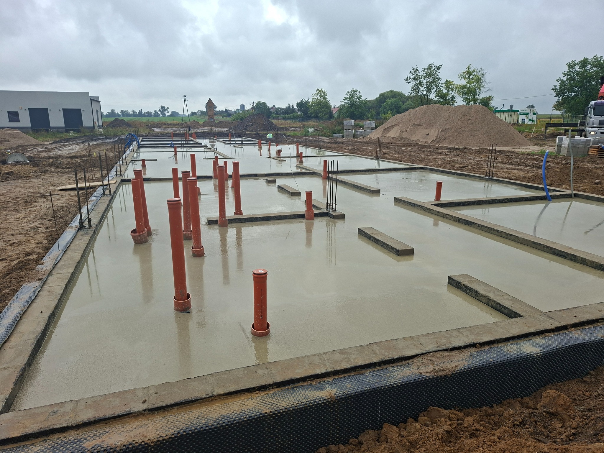 Zalany fundament z pionowymi rurami kanalizacyjnymi na budowie. Widoczne zbrojenia, budynek w tle i hałda piasku. Pochmurne niebo.