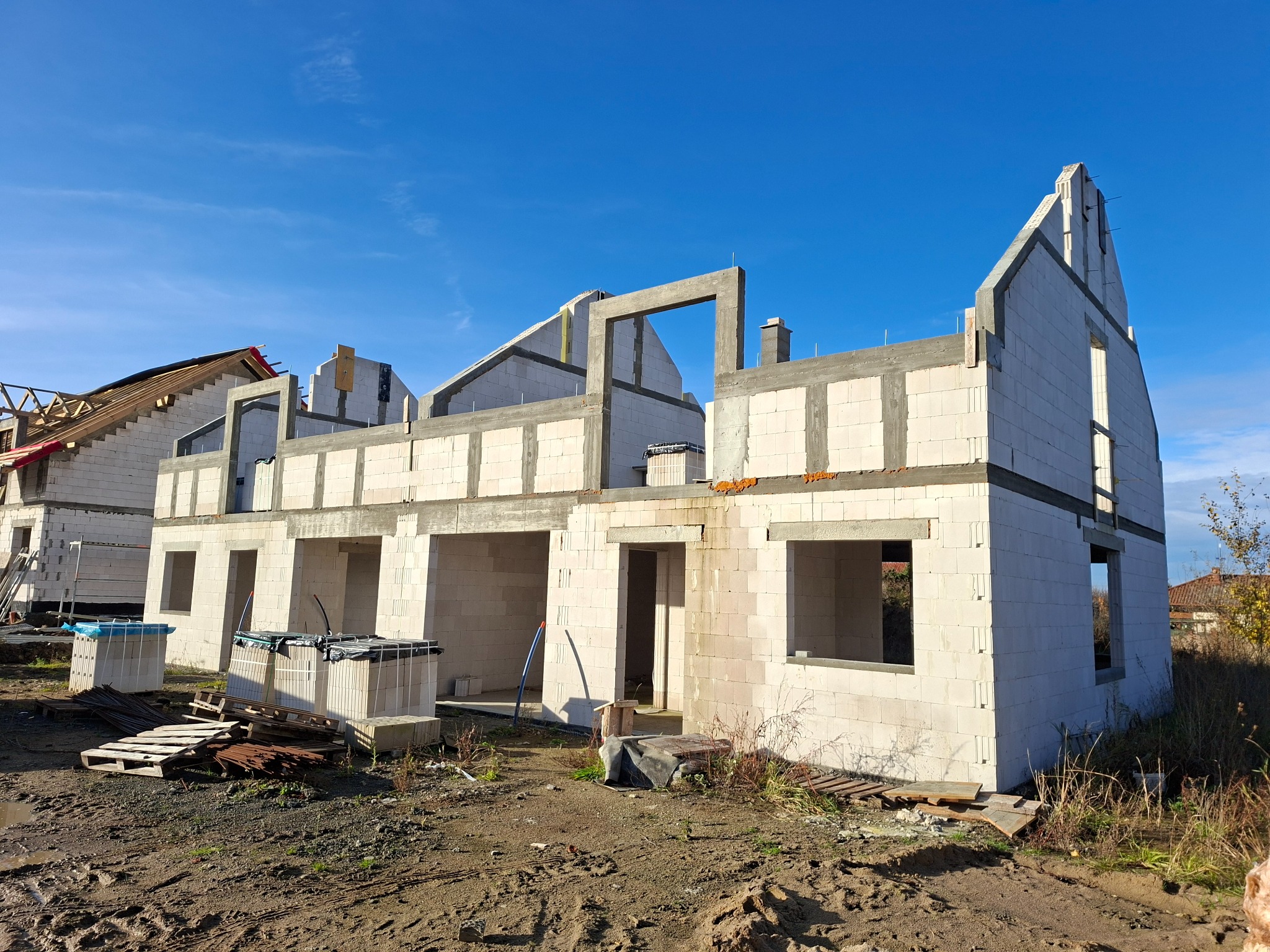 Wznoszenie surowego stanu domu z betonu komórkowego. Widoczne ściany, otwory okienne i drzwiowe. Częściowo wykonana więźba dachowa. Błękitne niebo w tle.