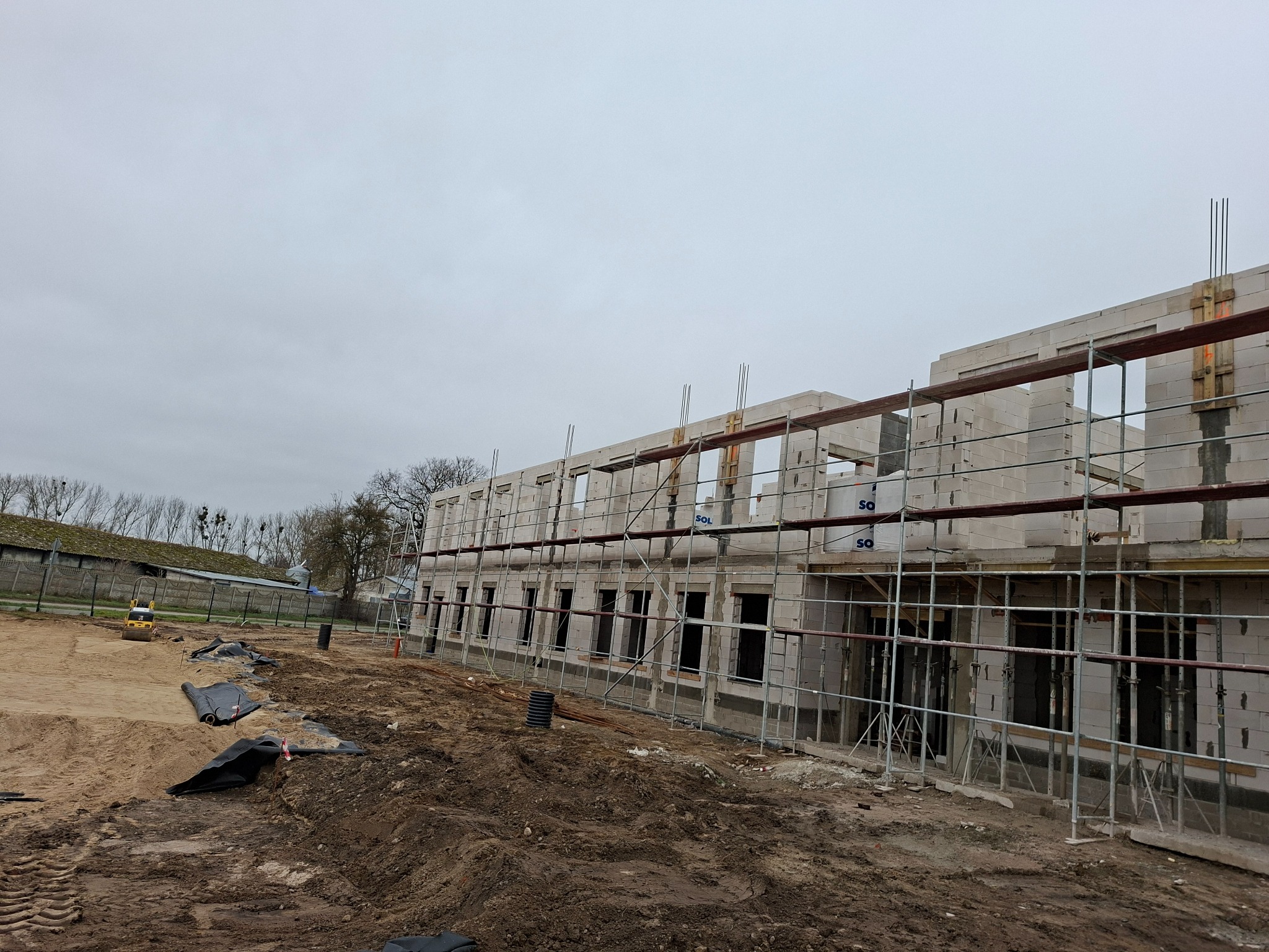 Wznoszenie dwukondygnacyjnego budynku z białych bloczków, widoczne rusztowania i teren budowy w pochmurny dzień. Etap murowania ścian.