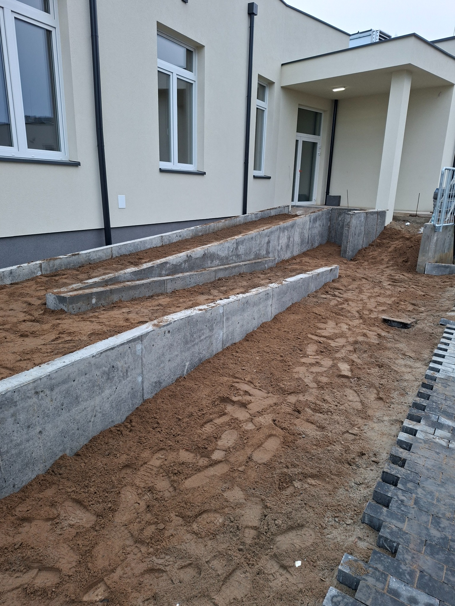 Betonowe rampy z piaskiem przed budynkiem, przygotowane pod układanie kostki brukowej. Widoczne fragmenty ułożonej kostki. Ujęcie z bliska, z ziemi.