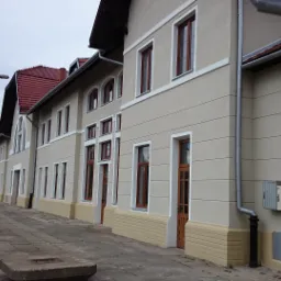 Elewacja budynku dworca kolejowego z beżową fasadą, czerwonymi ramami okien i drzwiami. Widoczna peron i elementy infrastruktury kolejowej.