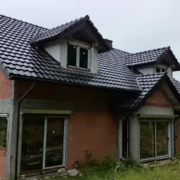 Dom w trakcie budowy z zamontowanymi oknami i roletami zewnętrznymi, widok z zewnątrz na tle zieleni i pochmurnego nieba.