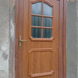 Brązowe drzwi wejściowe z szybą, wstawione w surowy otwór murarski. Widoczna złota klamka i górna szyba doświetlająca.