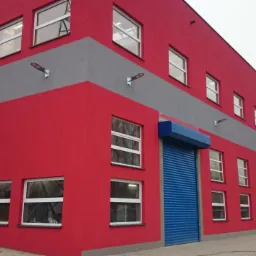 Czerwono-szary budynek z białymi oknami i niebieską bramą rolowaną. Widok z zewnątrz, oświetlenie uliczne i zaparkowany samochód w tle.